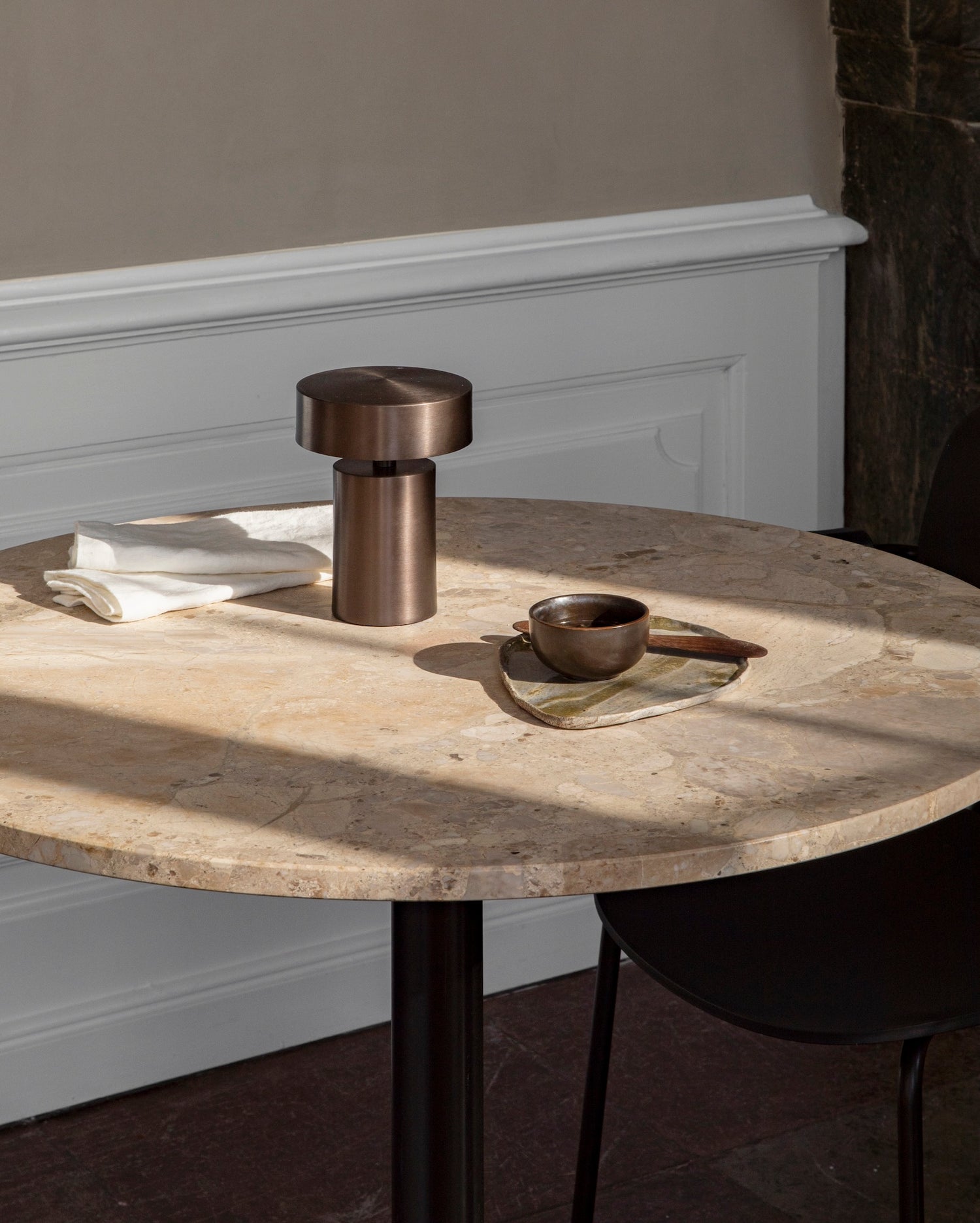 Close up of the harbour column dining table by audo copenhagen, black base with kunis breccia stone, plate and portable lamp on top.