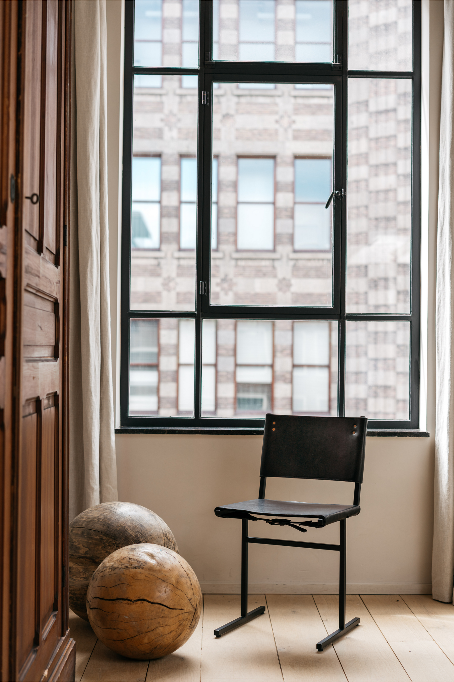 The Memento Chair, Black Frame by WDSTCK. the chair is in front of a large window with black frames. To the left is a classic wooden cabinet. and you can see 2 large accessory items on the floor. 