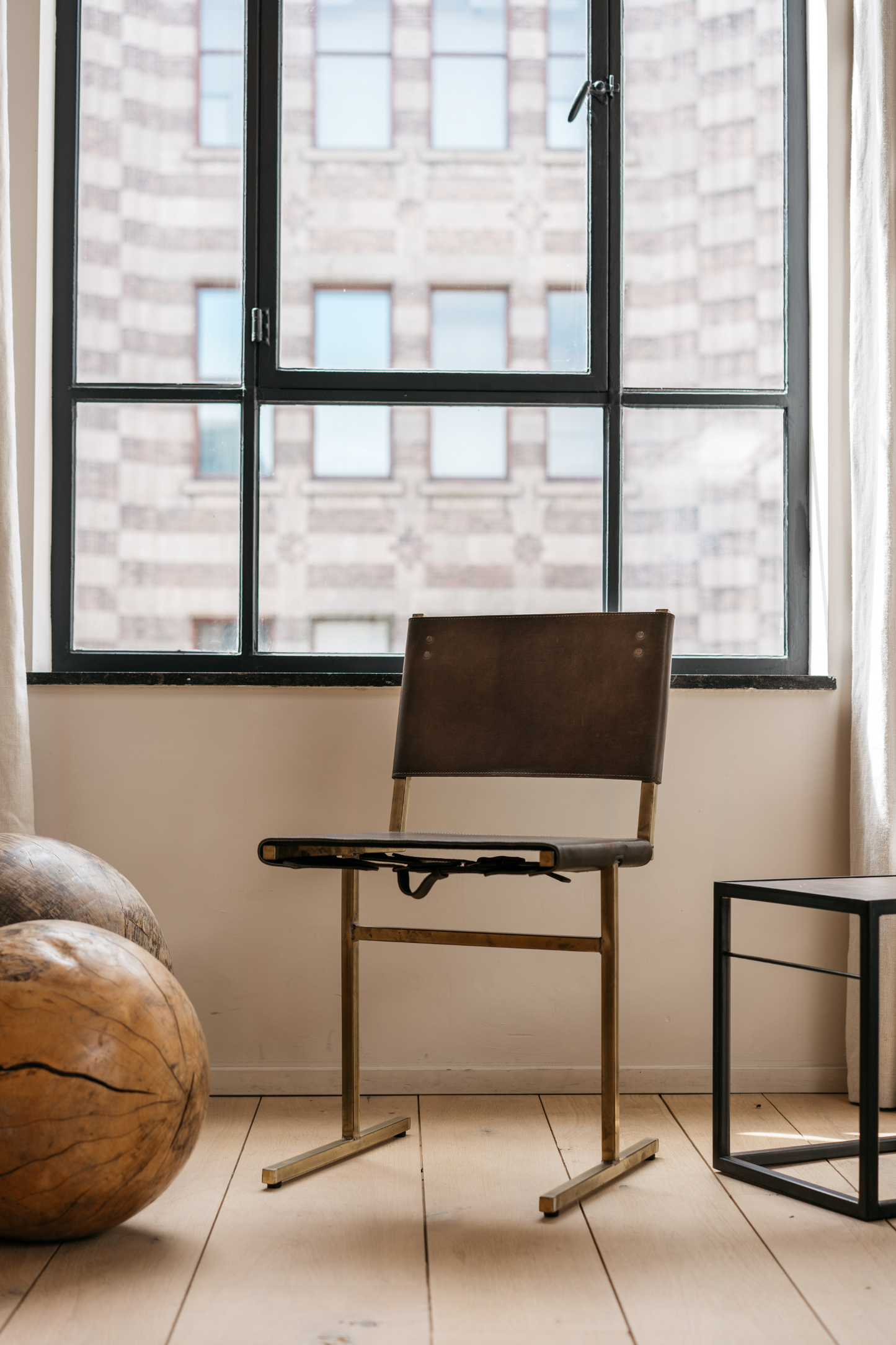 The Memento Chair, Brass Frame by WDSTCK. the chair is in front of a large window with black frames. To the left is a classic wooden cabinet. and you can see 2 large accessory items on the floor.