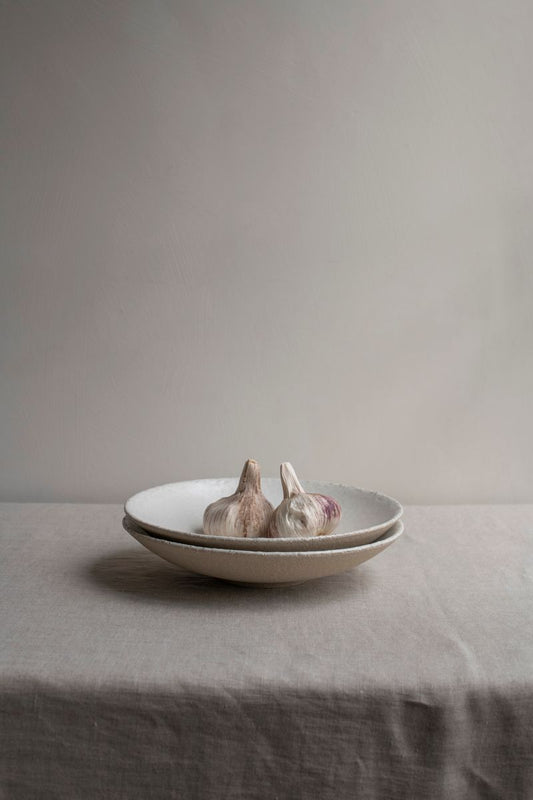 Two stacked Wabi Deep Plates by Jars Ceramistes. Set on white linen table cloth in front of white wall. Containing garlic in the plate.
