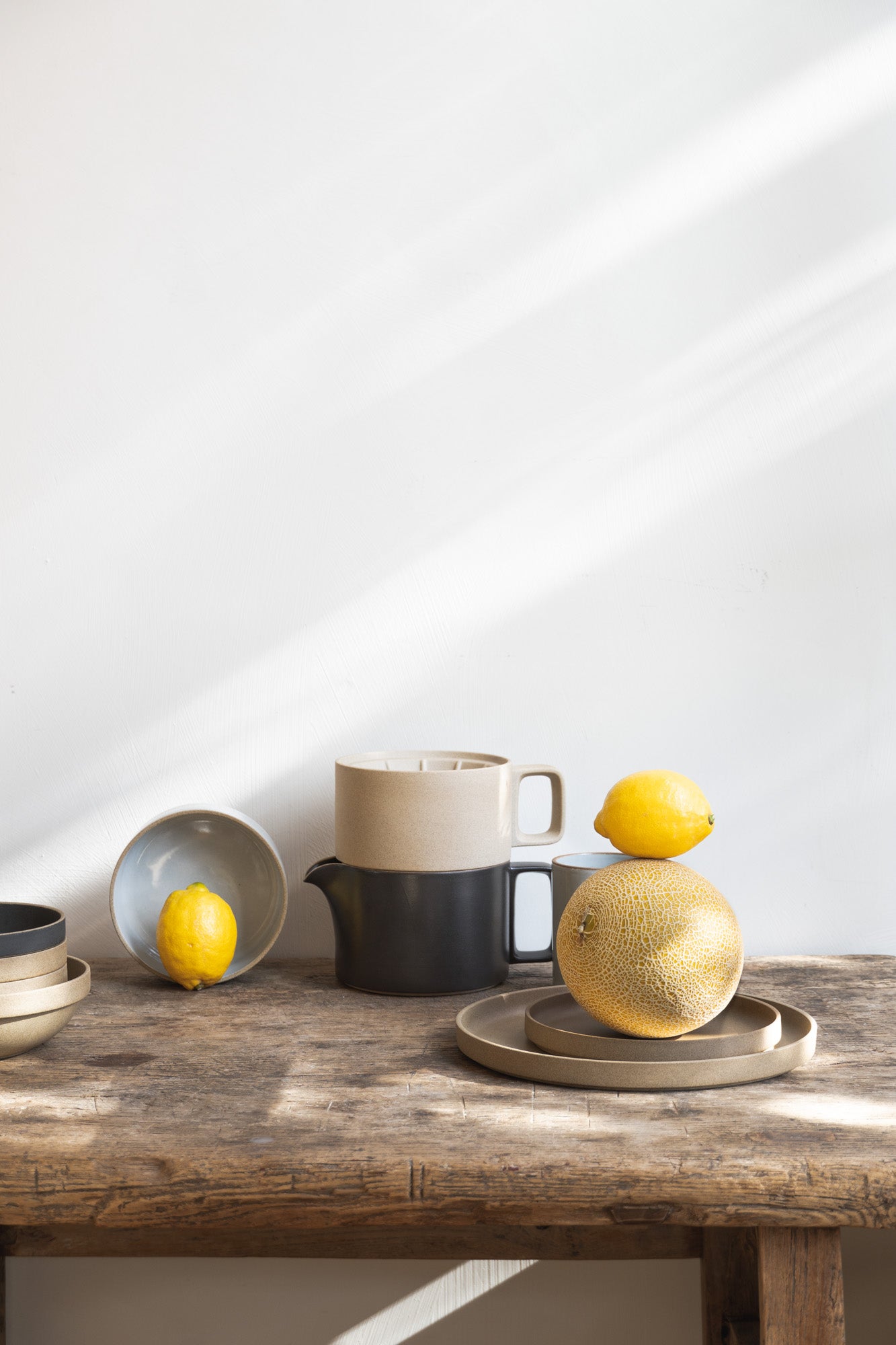 Rustic wooden table set with plates, mugs and coffee dripper. Accompanied by some lemons and a melon. All stackable and modular items by Hasami Porcelain.