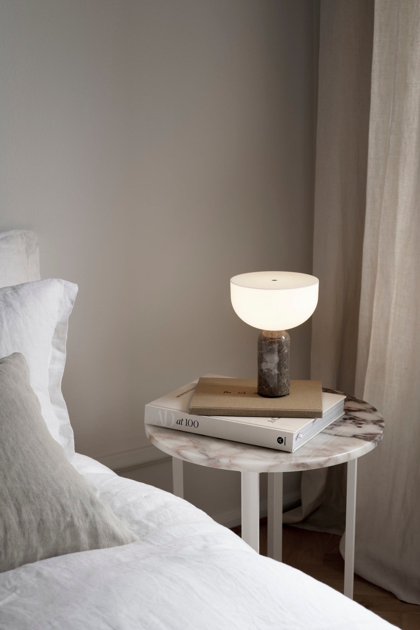 Kizu Portable Lamp Gris du Marais by New Works. Set on books on side table in neutral bedroom interior.