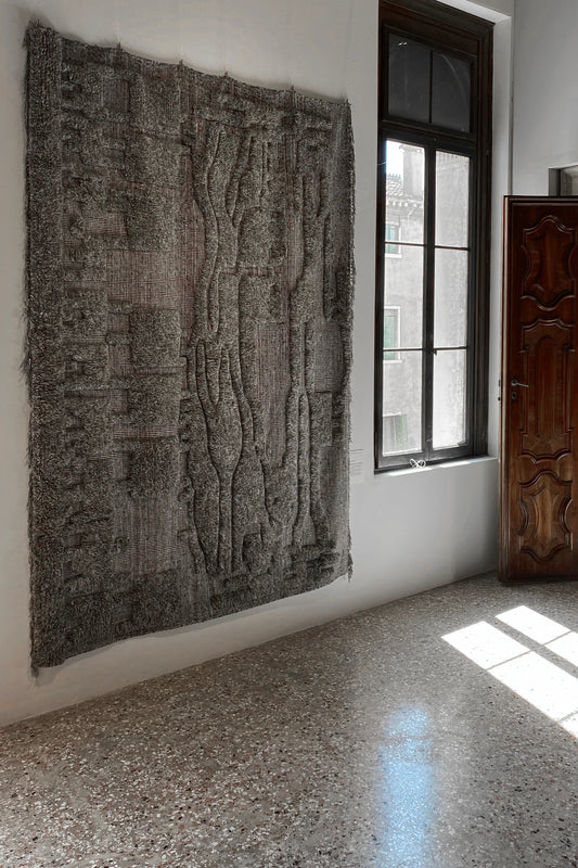 The grey PCB Rug by Zakaria Rugs hanging from a wall in a light interior room.